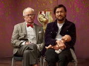 An older grey white man in a suit and glasses sitting next to a white man holding a doll with a cross section model of a pregnant belly behind them