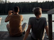 Two white men facing away from us into a lake with forest in the background. One of them is taking a picture.