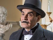 White man with wide moustache wearing a suit and top hat. In the background the bust of an ancient philosopher