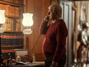 A white man in an brown jumper is talking on a landline phone. There is wood panellin on the walls and a bookshelf with old books. A display of full body medieval armour is on display in the background