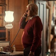 A white man in an brown jumper is talking on a landline phone. There is wood panellin on the walls and a bookshelf with old books. A display of full body medieval armour is on display in the background
