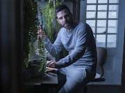 White man with short dark hair wearing a grey tracksuit sitting at a desk surrounded by plants. Behind him a window with bars.