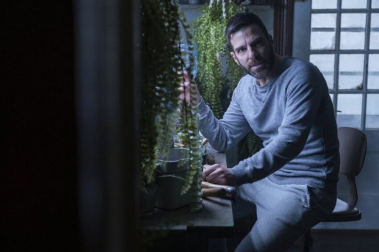 White man with short dark hair wearing a grey tracksuit sitting at a desk surrounded by plants. Behind him a window with bars.