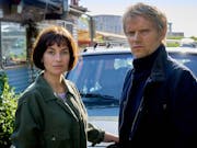 A white woman with short dark hair and a white blonde man wearing winter clothes stand in front of a car looking sternly towards the camera.