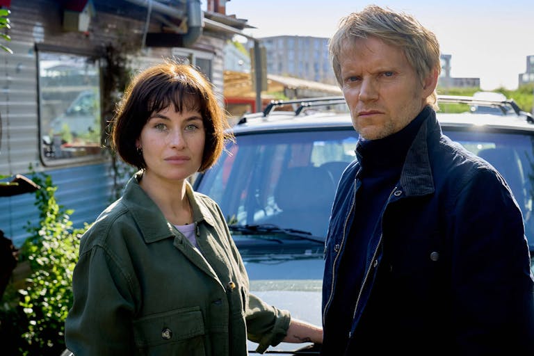 A white woman with short dark hair and a white blonde man wearing winter clothes stand in front of a car looking sternly towards the camera.