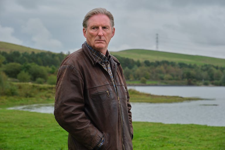 White man with short grey hair wearing a thick brown jacket looks sternly towards the camera. In the background a lake surrounded by green valley