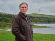 White man with short grey hair wearing a thick brown jacket looks sternly towards the camera. In the background a lake surrounded by green valley