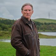 White man with short grey hair wearing a thick brown jacket looks sternly towards the camera. In the background a lake surrounded by green valley