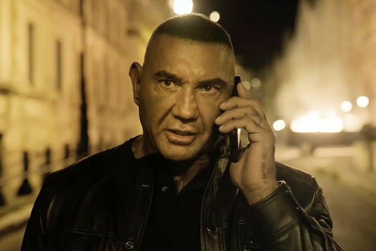 A man wearing a leather jacket is speaking on the phone in the middle of the street at night.