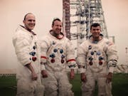 Three white man in space suits, smiling in front of a space launch system