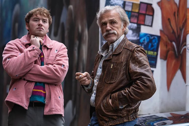 Two white men; a youger one in a pink jacket and an older one with a white hair and a wide moustache wearing a brown leather jacket standing in front of a graffitied wall observing something in the distance.