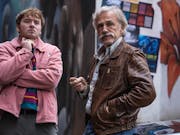 Two white men; a youger one in a pink jacket and an older one with a white hair and a wide moustache wearing a brown leather jacket standing in front of a graffitied wall observing something in the distance.