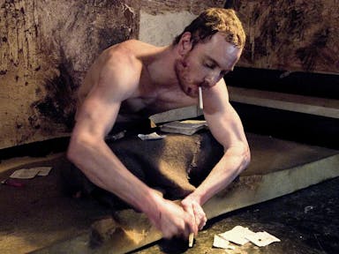 White man with short hair, sitting on a torn mattress on the floor, in a filthy room with newspaper clippings near him is smoking a cigarette whilst trying to cut something with his hands.