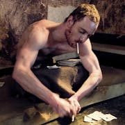 White man with short hair, sitting on a torn mattress on the floor, in a filthy room with newspaper clippings near him is smoking a cigarette whilst trying to cut something with his hands.