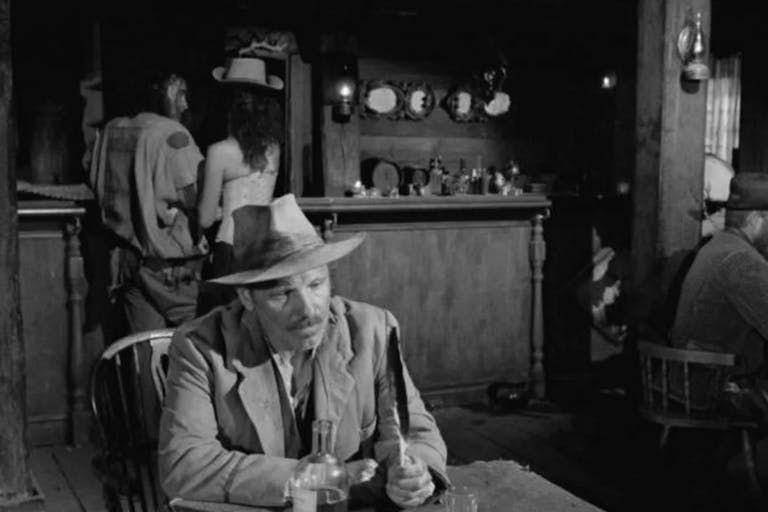 Black & white image of a white man wearing a hat sitting at a table holding a feather. In the background a wooden bar and a man and woman stand in front of it with their backs at us.