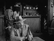 Black & white image of a white man wearing a hat sitting at a table holding a feather. In the background a wooden bar and a man and woman stand in front of it with their backs at us.
