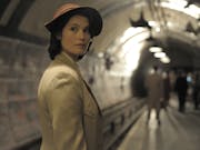 White woman in 40s clothing, wearing a hat is standing on a platform on the London Tube. In the background few more people standing or walking.