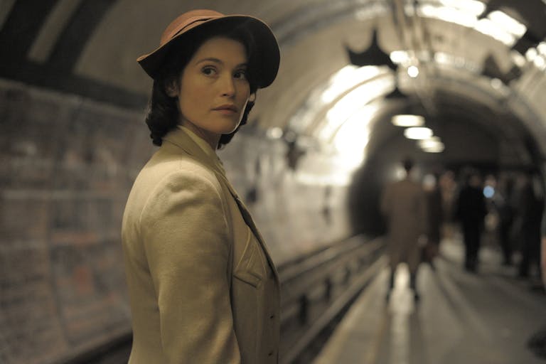 White woman in 40s clothing, wearing a hat is standing on a platform on the London Tube. In the background  few more people standing or walking.