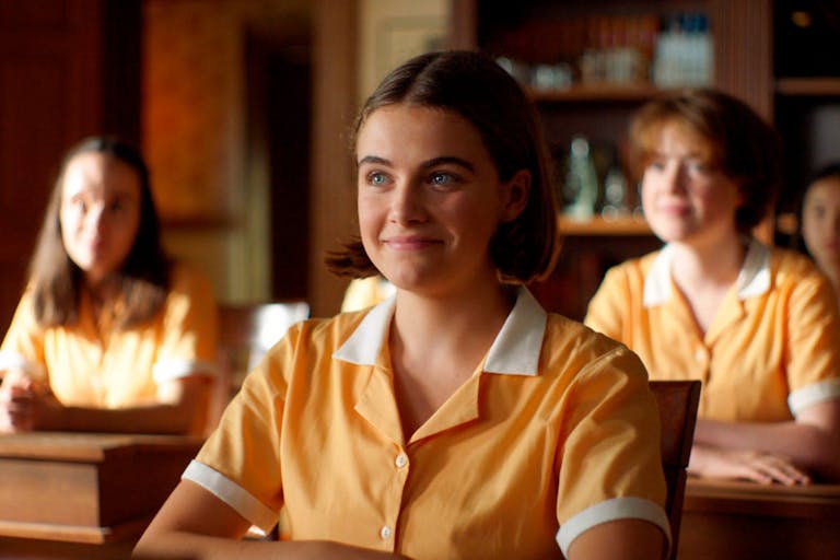 A young white girl in a yellow school uniform, sitting at a desk smiling. In the background more students sitting at their desks