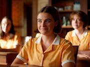 A young white girl in a yellow school uniform, sitting at a desk smiling. In the background more students sitting at their desks