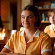 A young white girl in a yellow school uniform, sitting at a desk smiling. In the background more students sitting at their desks