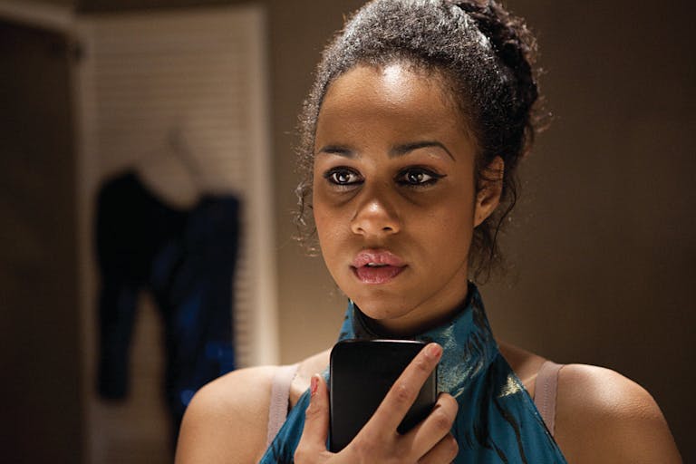 A black woman wearing a green satin dress is holding a phone in front of her looking pensive.