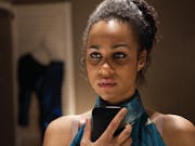 A black woman wearing a green satin dress is holding a phone in front of her looking pensive.