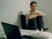 A white man with short hear sitting on a chair in the corner of room with his feet on the chair. An open laptop is on a bed with white sheets in the foreground.