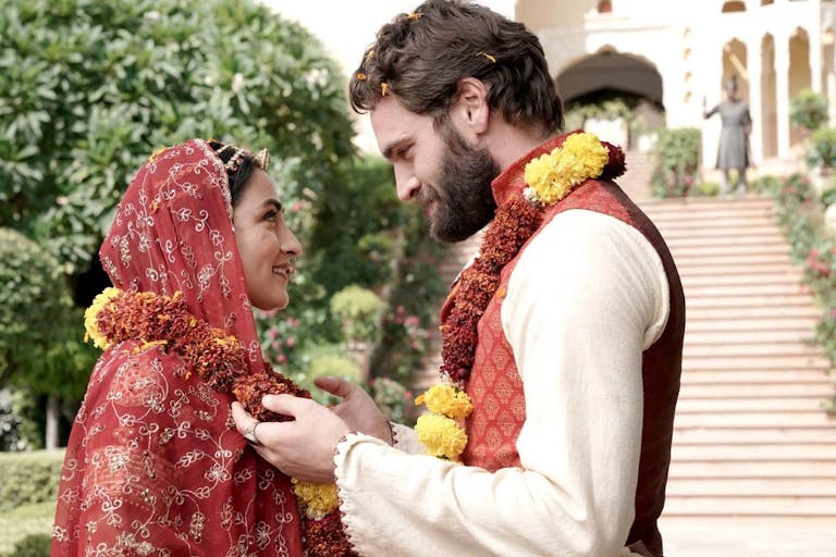 White man with beard and Asian woman in a sari, both wearing flowers around their necks looking at each other. In the background a grand building and gardens.