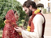 White man with beard and Asian woman in a sari, both wearing flowers around their necks looking at each other. In the background a grand building and gardens.