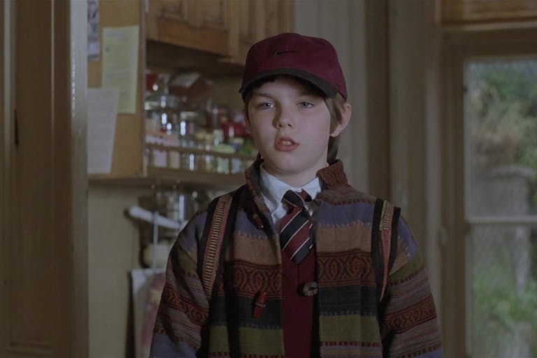 A young boy wearing a burgundy cap, a school uniform and a stripy wool jacket is looking curious. Behind him a kitchen cupboard and a kid's drawing of a flower on the wall.