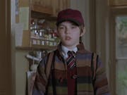A young boy wearing a burgundy cap, a school uniform and a stripy wool jacket is looking curious. Behind him a kitchen cupboard and a kid's drawing of a flower on the wall.