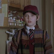 A young boy wearing a burgundy cap, a school uniform and a stripy wool jacket is looking curious. Behind him a kitchen cupboard and a kid's drawing of a flower on the wall.