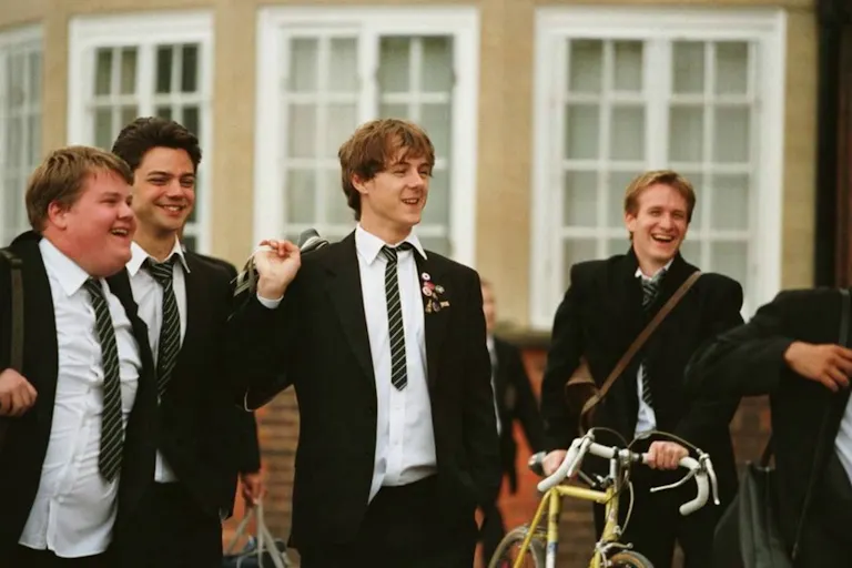 Five young men wearing suits are laughing at something off screen. Their shirts are untucked and they are holding backpacks over their shoulders. One of them is pushing a yellow bicycle along.