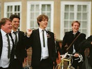 Five young men wearing suits are laughing at something off screen. Their shirts are untucked and they are holding backpacks over their shoulders. One of them is pushing a yellow bicycle along.