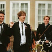 Five young men wearing suits are laughing at something off screen. Their shirts are untucked and they are holding backpacks over their shoulders. One of them is pushing a yellow bicycle along.