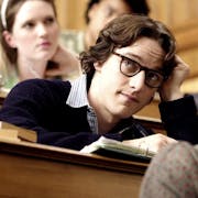 White young man with black thick rimmed glasses wearing a white shirt and black jumper on top is sitting at a university desk. Other students are at desks around hime