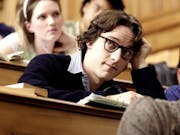 White young man with black thick rimmed glasses wearing a white shirt and black jumper on top is sitting at a university desk. Other students are at desks around hime