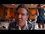 White man in a shiny suit looking closely and intensely at the camera. In the background restaurant tables with white table cloths and the back of a young woman with a black dress