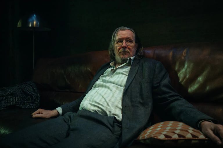 white man with greasy long hair wearing large glasses is sitting on a sofa. He is wearing a stripey white shirt and a grey suit.