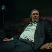 white man with greasy long hair wearing large glasses is sitting on a sofa. He is wearing a stripey white shirt and a grey suit.