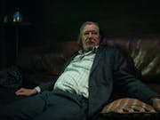 white man with greasy long hair wearing large glasses is sitting on a sofa. He is wearing a stripey white shirt and a grey suit.