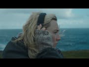 Blonde white woman, standing atop a cliff wears cut off grey gloves and is holding a pair of headphones over her head. In the background greys skies and rough seas.