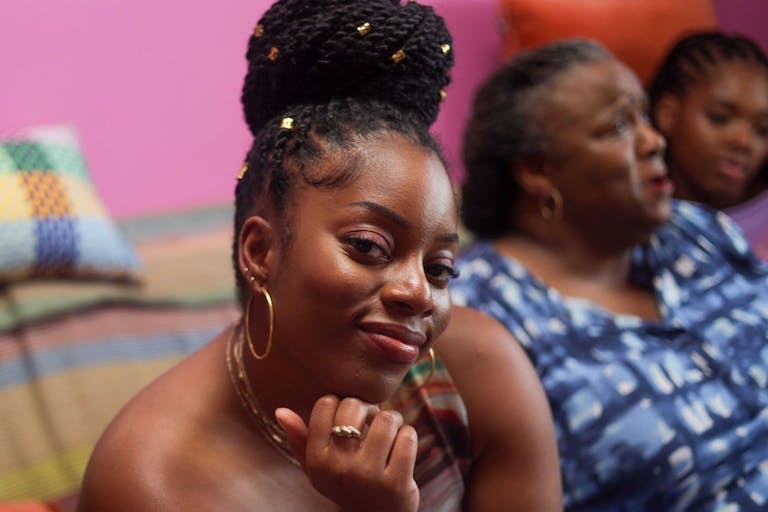 A young Black woman with long sleek braids pulled up into a large bun, sitting confidently and radiantly on the sofa next to an older Black woman, and a young Black girl