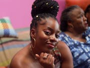 A young Black woman with long sleek braids pulled up into a large bun, sitting confidently and radiantly on the sofa next to an older Black woman, and a young Black girl
