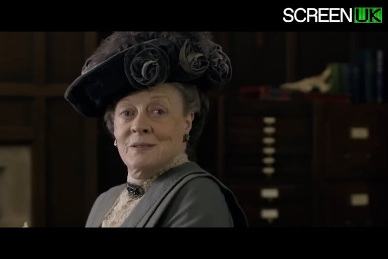 An old white woman in elegant period clothes and ornate hat looking happy and regal