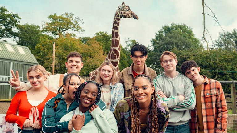 A varied group of teens smiling and posing in front of a giraffe