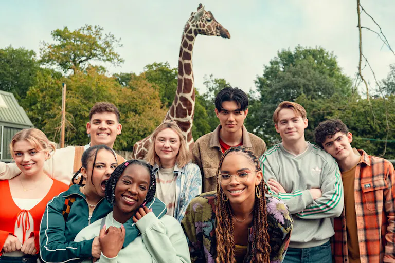 A varied group of teens smiling and posing in front of a giraffe