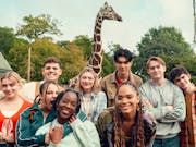 A varied group of teens smiling and posing in front of a giraffe