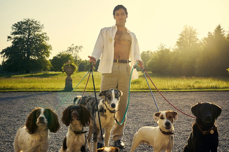 A white man with open button down shirt holding the leads of 5 dogs, standing in a country estate garden